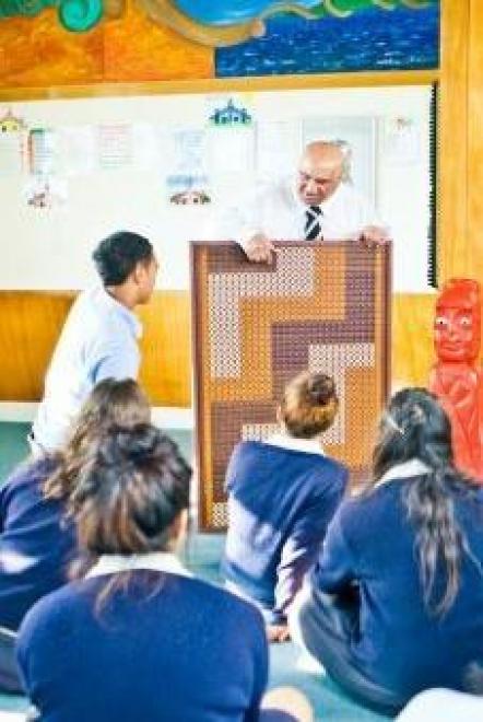 Older man holds a tukutuku panel. He is engaged with the school student who is looking at the panel. There are five other students in school uniform sitting cross legged on the floor facing the man, the student and the tukutuku panel. They are facing away from the camera.
