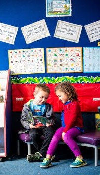 Two young students sharing a book in a classroom setting. There are alphabet phonics cards on the wall behind them.