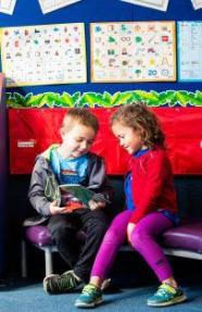 Two children sitting sharing a book under phonics alphabet cards on the wall behind them.